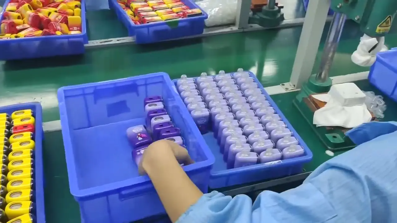 工場の組立ライン。青い袖を着た作業員が紫色のボトルを青いビンに入れます。近くの緑の生産テーブルには、透明なキャップが付いたグレーの小さなボトルが整然と並べられたトレイがあり、背景にはカラフルな製品が詰まった他の青いビンがあります。.